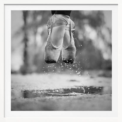 A close-up, black and white photograph of a ballerina's feet in worn pointe shoes, captured in mid-air above a puddle, with water splashing upwards. The motion is frozen, highlighting the contrast between grace and a raw outdoor element. The image is presented in a light wood frame. Poster