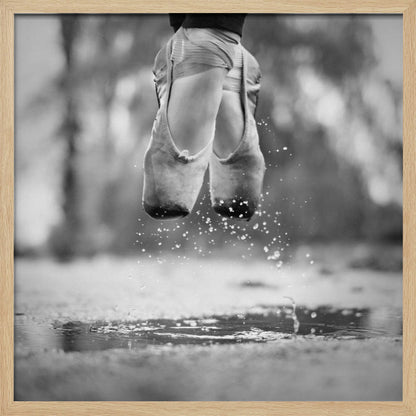 A close-up, black and white photograph of a ballerina's feet in worn pointe shoes, captured in mid-air above a puddle, with water splashing upwards. The motion is frozen, highlighting the contrast between grace and a raw outdoor element. The image is presented in a light wood frame. Poster