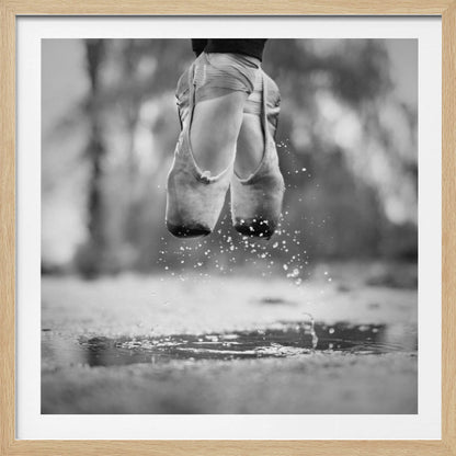 A close-up, black and white photograph of a ballerina's feet in worn pointe shoes, captured in mid-air above a puddle, with water splashing upwards. The motion is frozen, highlighting the contrast between grace and a raw outdoor element. The image is presented in a light wood frame. Poster