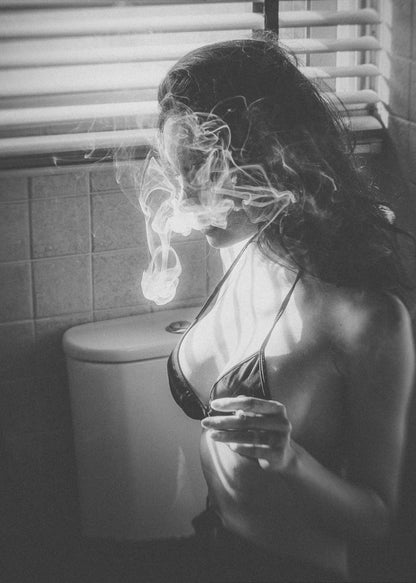 A black and white photograph of a woman in a bikini top smoking a cigarette in a bathroom. Light from a window with blinds creates dramatic shadows as a thick cloud of smoke obscures her face. Poster
