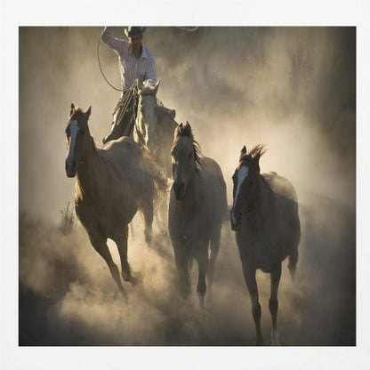 A dramatic, backlit photograph of a cowboy on horseback herding a small group of horses, kicking up a large cloud of dust as they gallop through a rustic landscape at sunrise or sunset. Poster