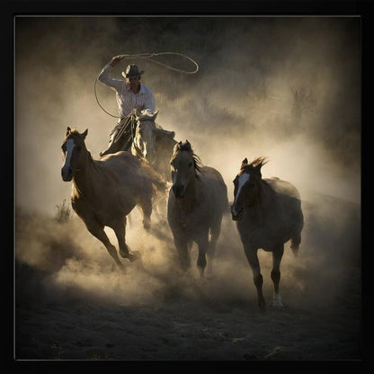A dramatic, backlit photograph of a cowboy on horseback herding a small group of horses, kicking up a large cloud of dust as they gallop through a rustic landscape at sunrise or sunset. Poster