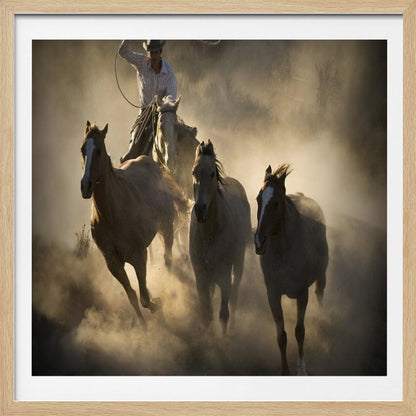 A dramatic, backlit photograph of a cowboy on horseback herding a small group of horses, kicking up a large cloud of dust as they gallop through a rustic landscape at sunrise or sunset. Poster