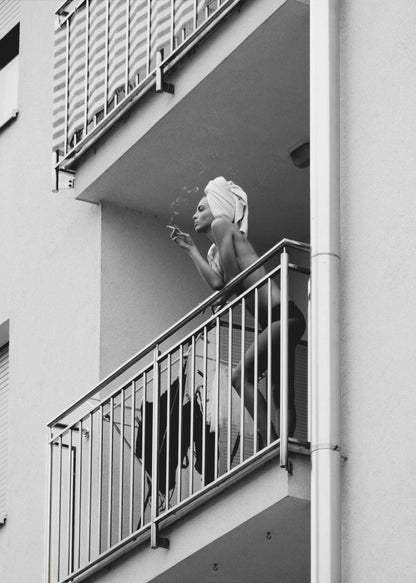A black and white photo of a woman with a towel on her head, smoking a cigarette on an apartment balcony. The low-angle shot captures her leaning on the metal railing, with smoke drifting from the cigarette. Print