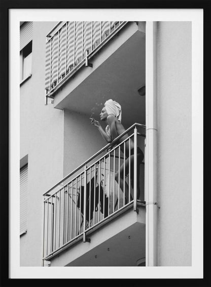 A black and white photo of a woman with a towel on her head, smoking a cigarette on an apartment balcony. The low-angle shot captures her leaning on the metal railing, with smoke drifting from the cigarette. Print
