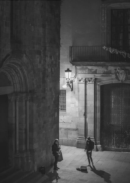 A high-angle, black and white photograph of a street musician playing a saxophone for a lone listener in a narrow stone alley at night. The scene is illuminated by an ornate streetlamp, casting long shadows on the cobblestone ground. Wall Art