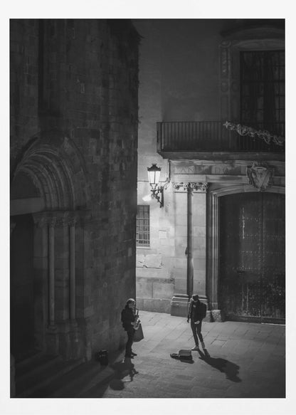 A high-angle, black and white photograph of a street musician playing a saxophone for a lone listener in a narrow stone alley at night. The scene is illuminated by an ornate streetlamp, casting long shadows on the cobblestone ground. Wall Art