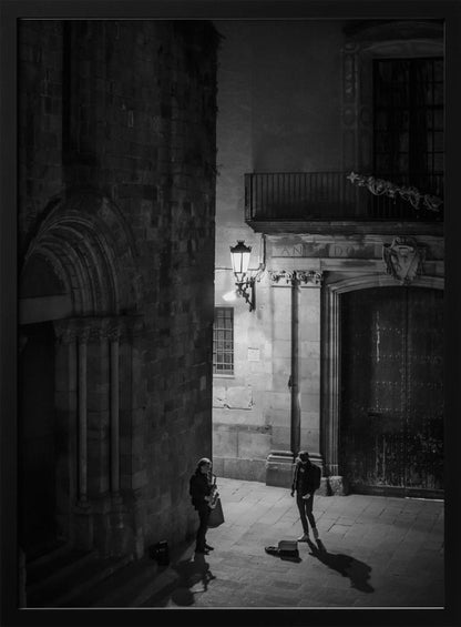 A high-angle, black and white photograph of a street musician playing a saxophone for a lone listener in a narrow stone alley at night. The scene is illuminated by an ornate streetlamp, casting long shadows on the cobblestone ground. Wall Art