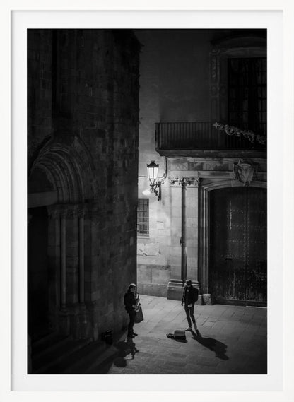 A high-angle, black and white photograph of a street musician playing a saxophone for a lone listener in a narrow stone alley at night. The scene is illuminated by an ornate streetlamp, casting long shadows on the cobblestone ground. Wall Art