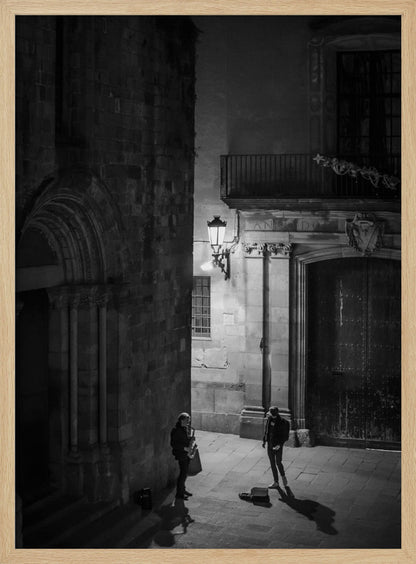 A high-angle, black and white photograph of a street musician playing a saxophone for a lone listener in a narrow stone alley at night. The scene is illuminated by an ornate streetlamp, casting long shadows on the cobblestone ground. Wall Art