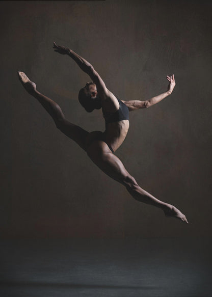 A dramatic, low-key studio photograph of a muscular female dancer captured mid-leap. She is in a full split, arching her back with her arms extended gracefully. The lighting accentuates her toned physique against a dark, textured brown background. Poster