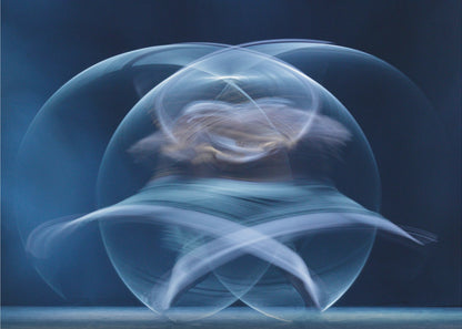 An abstract long-exposure photograph of a spinning dancer, creating ethereal, intersecting trails of blue and white light that form a glowing, orb-like shape against a deep blue background. The image is presented within a silver frame. Poster