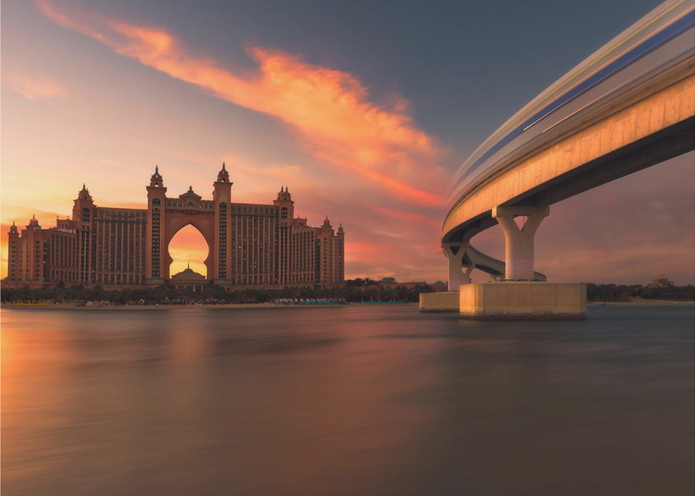 A scenic view of the Atlantis The Palm hotel in Dubai at sunset, with its grand architecture silhouetted against a vibrant orange and pink sky. A modern monorail track curves over the water on the right, with a blurred train indicating motion. The calm water in the foreground reflects the warm colors of the dusk sky. Decor
