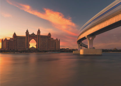 A scenic view of the Atlantis The Palm hotel in Dubai at sunset, with its grand architecture silhouetted against a vibrant orange and pink sky. A modern monorail track curves over the water on the right, with a blurred train indicating motion. The calm water in the foreground reflects the warm colors of the dusk sky. Decor