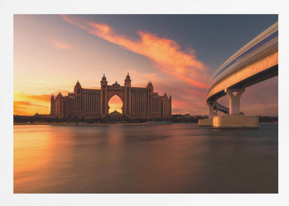 A scenic view of the Atlantis The Palm hotel in Dubai at sunset, with its grand architecture silhouetted against a vibrant orange and pink sky. A modern monorail track curves over the water on the right, with a blurred train indicating motion. The calm water in the foreground reflects the warm colors of the dusk sky. Decor