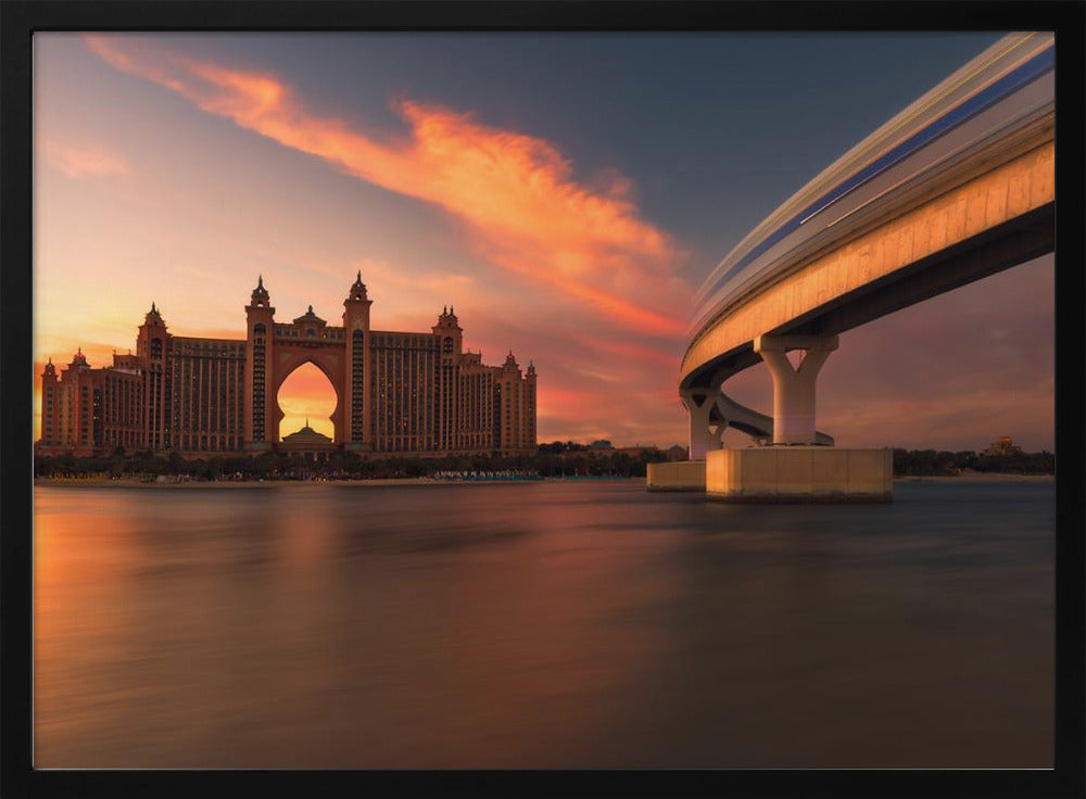 A scenic view of the Atlantis The Palm hotel in Dubai at sunset, with its grand architecture silhouetted against a vibrant orange and pink sky. A modern monorail track curves over the water on the right, with a blurred train indicating motion. The calm water in the foreground reflects the warm colors of the dusk sky. Decor