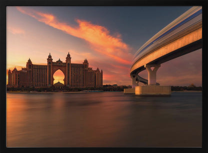 A scenic view of the Atlantis The Palm hotel in Dubai at sunset, with its grand architecture silhouetted against a vibrant orange and pink sky. A modern monorail track curves over the water on the right, with a blurred train indicating motion. The calm water in the foreground reflects the warm colors of the dusk sky. Decor