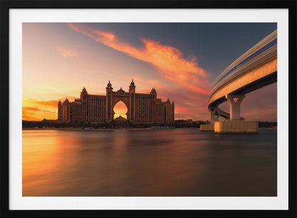 A scenic view of the Atlantis The Palm hotel in Dubai at sunset, with its grand architecture silhouetted against a vibrant orange and pink sky. A modern monorail track curves over the water on the right, with a blurred train indicating motion. The calm water in the foreground reflects the warm colors of the dusk sky. Decor
