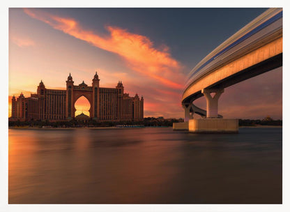 A scenic view of the Atlantis The Palm hotel in Dubai at sunset, with its grand architecture silhouetted against a vibrant orange and pink sky. A modern monorail track curves over the water on the right, with a blurred train indicating motion. The calm water in the foreground reflects the warm colors of the dusk sky. Decor