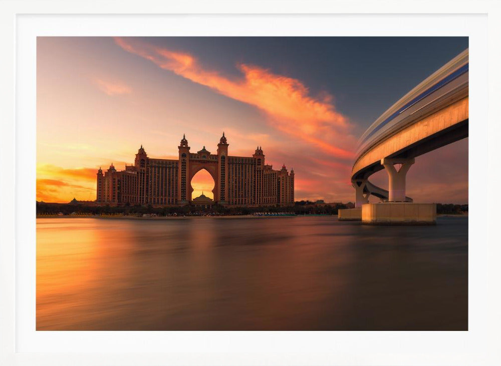 A scenic view of the Atlantis The Palm hotel in Dubai at sunset, with its grand architecture silhouetted against a vibrant orange and pink sky. A modern monorail track curves over the water on the right, with a blurred train indicating motion. The calm water in the foreground reflects the warm colors of the dusk sky. Decor