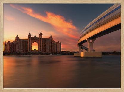 A scenic view of the Atlantis The Palm hotel in Dubai at sunset, with its grand architecture silhouetted against a vibrant orange and pink sky. A modern monorail track curves over the water on the right, with a blurred train indicating motion. The calm water in the foreground reflects the warm colors of the dusk sky. Decor