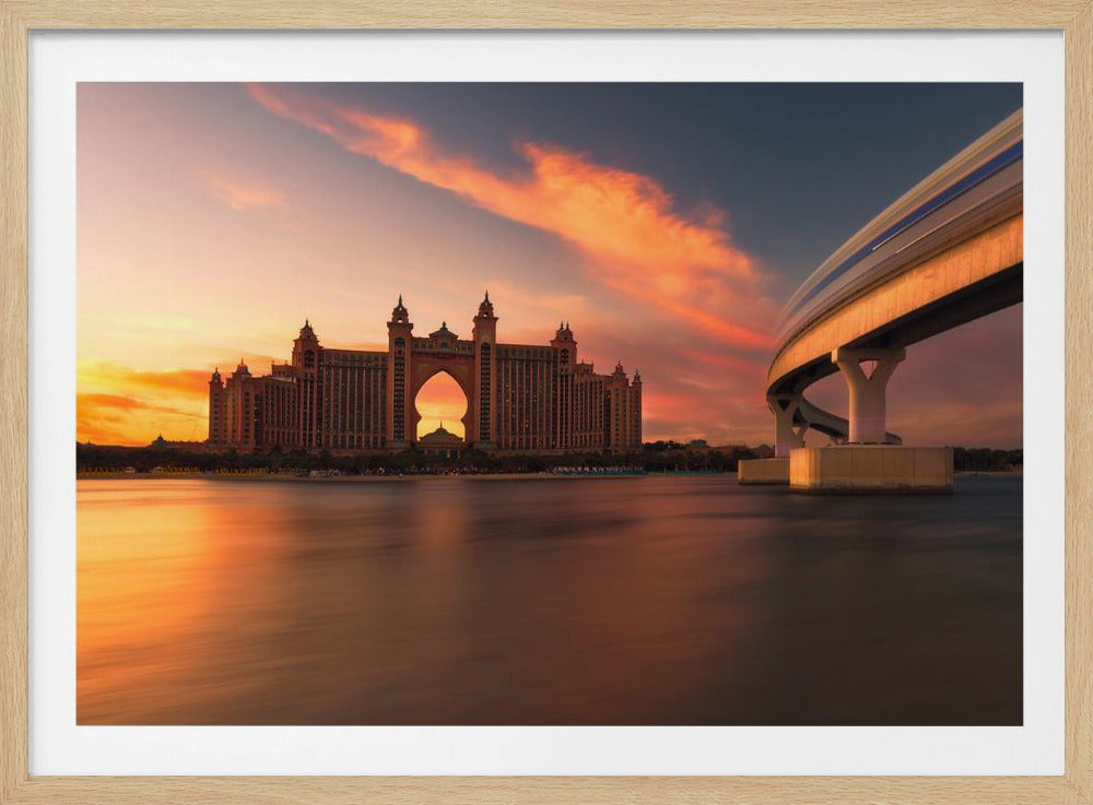 A scenic view of the Atlantis The Palm hotel in Dubai at sunset, with its grand architecture silhouetted against a vibrant orange and pink sky. A modern monorail track curves over the water on the right, with a blurred train indicating motion. The calm water in the foreground reflects the warm colors of the dusk sky. Decor