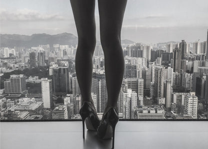A framed black and white photograph showing the back of a woman's legs and high heels as she stands before a large window, looking out over a vast, dense cityscape with distant mountains under a cloudy sky. Artwork