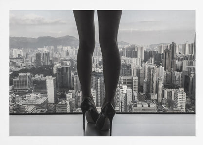 A framed black and white photograph showing the back of a woman's legs and high heels as she stands before a large window, looking out over a vast, dense cityscape with distant mountains under a cloudy sky. Artwork