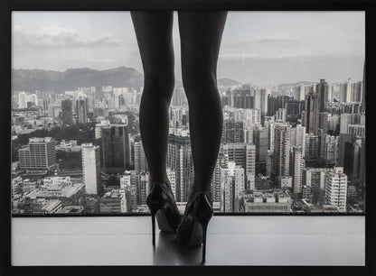 A framed black and white photograph showing the back of a woman's legs and high heels as she stands before a large window, looking out over a vast, dense cityscape with distant mountains under a cloudy sky. Artwork