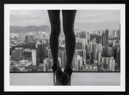A framed black and white photograph showing the back of a woman's legs and high heels as she stands before a large window, looking out over a vast, dense cityscape with distant mountains under a cloudy sky. Artwork