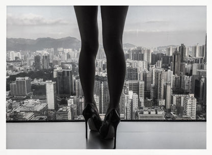 A framed black and white photograph showing the back of a woman's legs and high heels as she stands before a large window, looking out over a vast, dense cityscape with distant mountains under a cloudy sky. Artwork