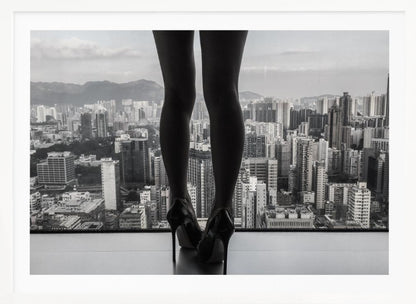 A framed black and white photograph showing the back of a woman's legs and high heels as she stands before a large window, looking out over a vast, dense cityscape with distant mountains under a cloudy sky. Artwork