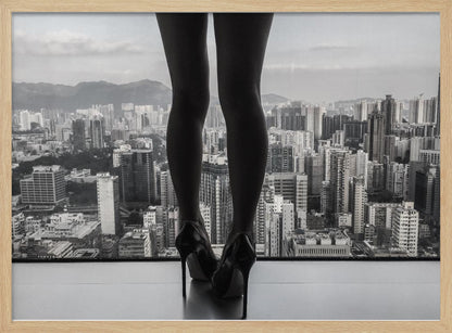 A framed black and white photograph showing the back of a woman's legs and high heels as she stands before a large window, looking out over a vast, dense cityscape with distant mountains under a cloudy sky. Artwork