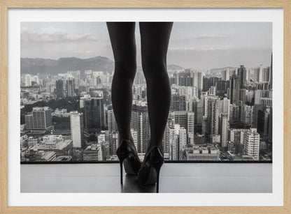 A framed black and white photograph showing the back of a woman's legs and high heels as she stands before a large window, looking out over a vast, dense cityscape with distant mountains under a cloudy sky. Artwork