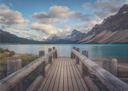 A wooden pier leads out onto a vibrant turquoise glacial lake, surrounded by towering, snow-dusted mountains under a dramatic cloudy sky. The image is presented in a silver frame. Artwork