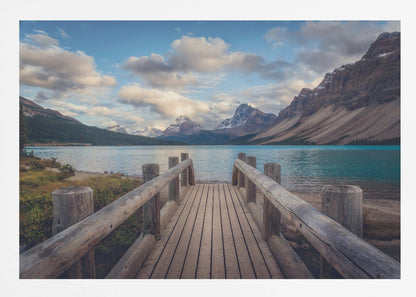 A wooden pier leads out onto a vibrant turquoise glacial lake, surrounded by towering, snow-dusted mountains under a dramatic cloudy sky. The image is presented in a silver frame. Artwork