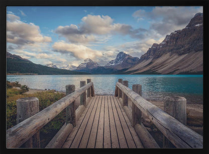 A wooden pier leads out onto a vibrant turquoise glacial lake, surrounded by towering, snow-dusted mountains under a dramatic cloudy sky. The image is presented in a silver frame. Artwork