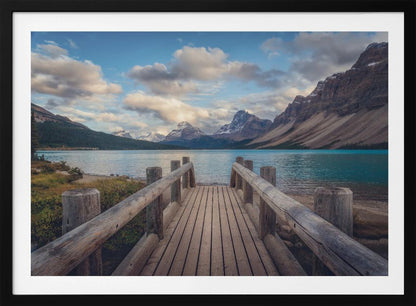 A wooden pier leads out onto a vibrant turquoise glacial lake, surrounded by towering, snow-dusted mountains under a dramatic cloudy sky. The image is presented in a silver frame. Artwork