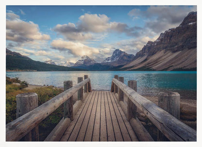 A wooden pier leads out onto a vibrant turquoise glacial lake, surrounded by towering, snow-dusted mountains under a dramatic cloudy sky. The image is presented in a silver frame. Artwork
