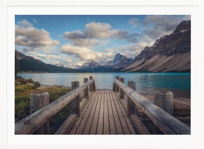 A wooden pier leads out onto a vibrant turquoise glacial lake, surrounded by towering, snow-dusted mountains under a dramatic cloudy sky. The image is presented in a silver frame. Artwork