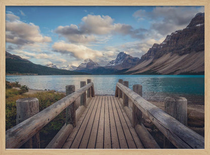 A wooden pier leads out onto a vibrant turquoise glacial lake, surrounded by towering, snow-dusted mountains under a dramatic cloudy sky. The image is presented in a silver frame. Artwork