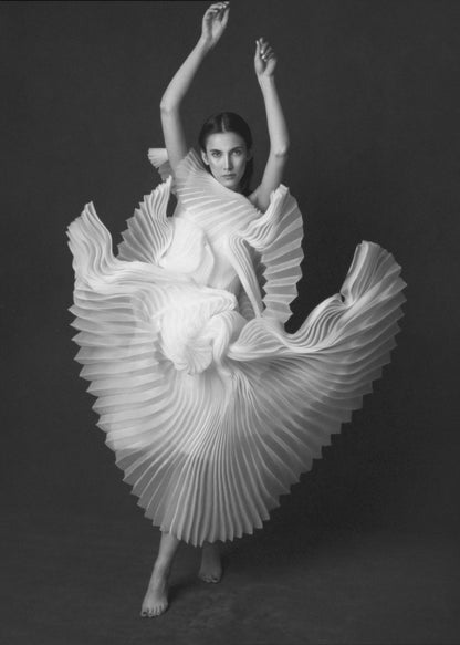 A framed, black and white photograph of a woman in a dramatic pose, wearing a voluminous white pleated dress. Her arms are raised above her head as the dress fans out around her in a sculptural, swirling motion. She looks directly at the camera against a solid dark gray background. Decor