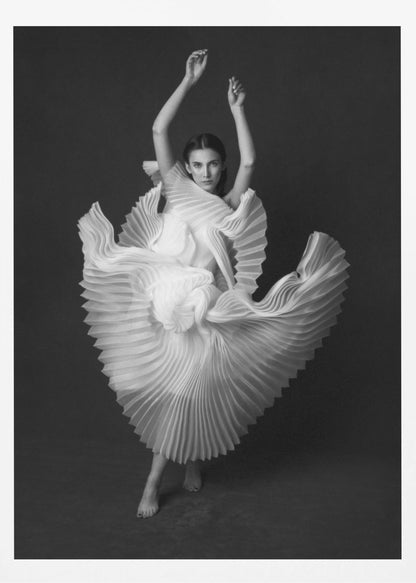 A framed, black and white photograph of a woman in a dramatic pose, wearing a voluminous white pleated dress. Her arms are raised above her head as the dress fans out around her in a sculptural, swirling motion. She looks directly at the camera against a solid dark gray background. Decor