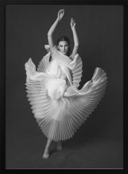 A framed, black and white photograph of a woman in a dramatic pose, wearing a voluminous white pleated dress. Her arms are raised above her head as the dress fans out around her in a sculptural, swirling motion. She looks directly at the camera against a solid dark gray background. Decor