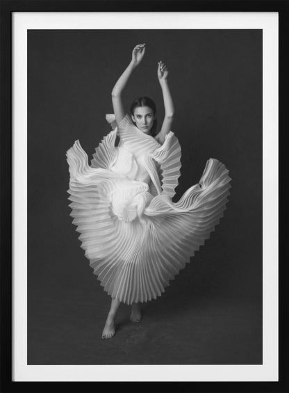 A framed, black and white photograph of a woman in a dramatic pose, wearing a voluminous white pleated dress. Her arms are raised above her head as the dress fans out around her in a sculptural, swirling motion. She looks directly at the camera against a solid dark gray background. Decor