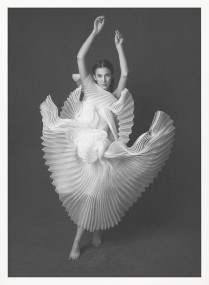 A framed, black and white photograph of a woman in a dramatic pose, wearing a voluminous white pleated dress. Her arms are raised above her head as the dress fans out around her in a sculptural, swirling motion. She looks directly at the camera against a solid dark gray background. Decor