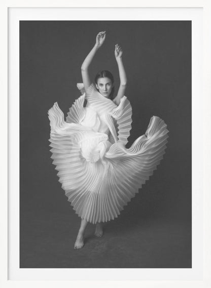 A framed, black and white photograph of a woman in a dramatic pose, wearing a voluminous white pleated dress. Her arms are raised above her head as the dress fans out around her in a sculptural, swirling motion. She looks directly at the camera against a solid dark gray background. Decor