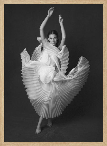 A framed, black and white photograph of a woman in a dramatic pose, wearing a voluminous white pleated dress. Her arms are raised above her head as the dress fans out around her in a sculptural, swirling motion. She looks directly at the camera against a solid dark gray background. Decor