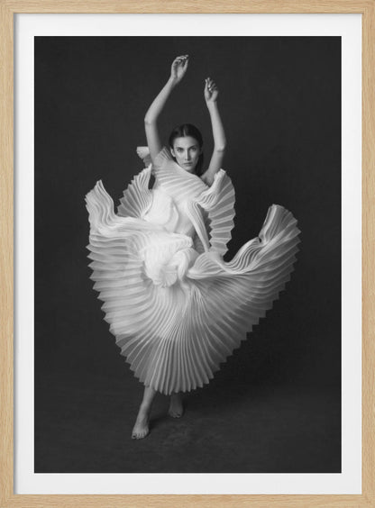 A framed, black and white photograph of a woman in a dramatic pose, wearing a voluminous white pleated dress. Her arms are raised above her head as the dress fans out around her in a sculptural, swirling motion. She looks directly at the camera against a solid dark gray background. Decor