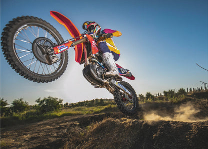 A dynamic, low-angle shot of a motocross rider in mid-air, jumping over a dirt mound. The rider is on a red, white, and blue dirt bike, wearing colorful racing gear and a helmet. The bike's front wheel is high in the air against a clear blue sky, while the back wheel is just clearing the jump, kicking up a cloud of dust. The background features a green, rural landscape. Wall Art